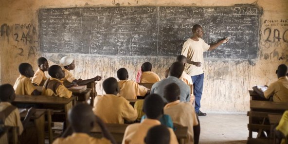 Un enseignant debout face à un tableau noir écrit des informations devant des élèves assis en uniforme dans une salle de classe simple