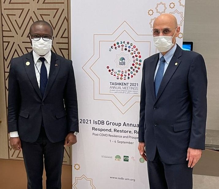 Deux hommes en costume sombre et masque chirurgical posent devant une bannière d'une conférence internationale