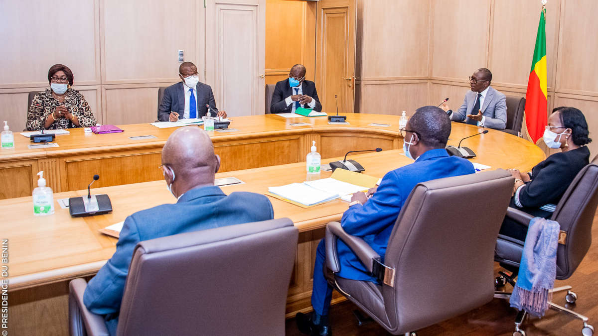Sept personnes assises autour d’une table de réunion avec drapeau et gel hydroalcoolique visibles