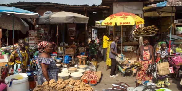 Marché animé avec des vendeuses et acheteurs autour de divers produits alimentaires en plein air sous parasols.