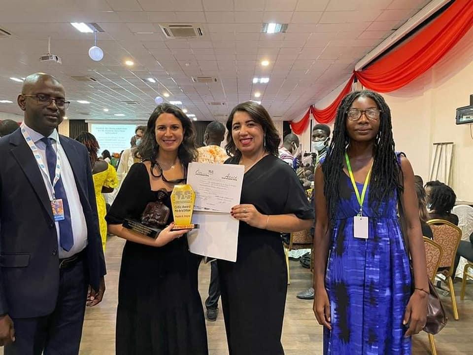 Quatre personnes debout, deux femmes au centre tiennent un trophée et un document, salle de réunion animée en arrière-plan