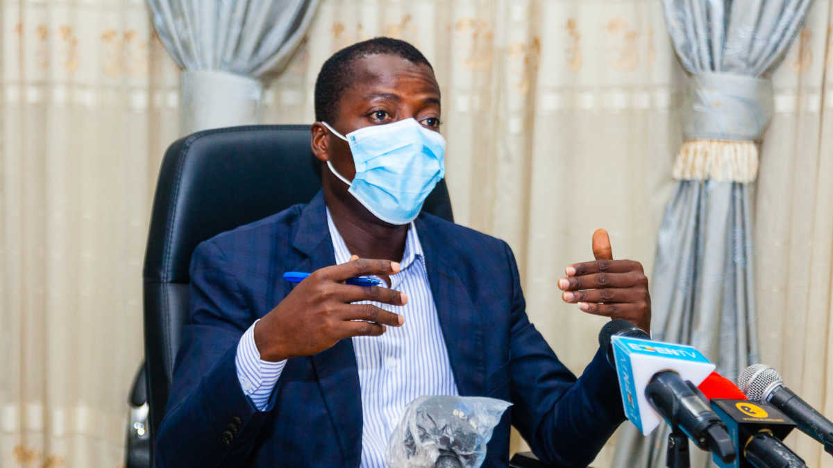 Un homme portant un masque parle en gestes derrière un micro avec rideaux clairs en arrière-plan