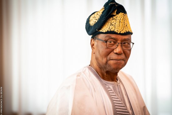 Homme âgé portant un boubou traditionnel blanc et un chapeau orné de motifs dorés souriant légèrement face à l'appareil