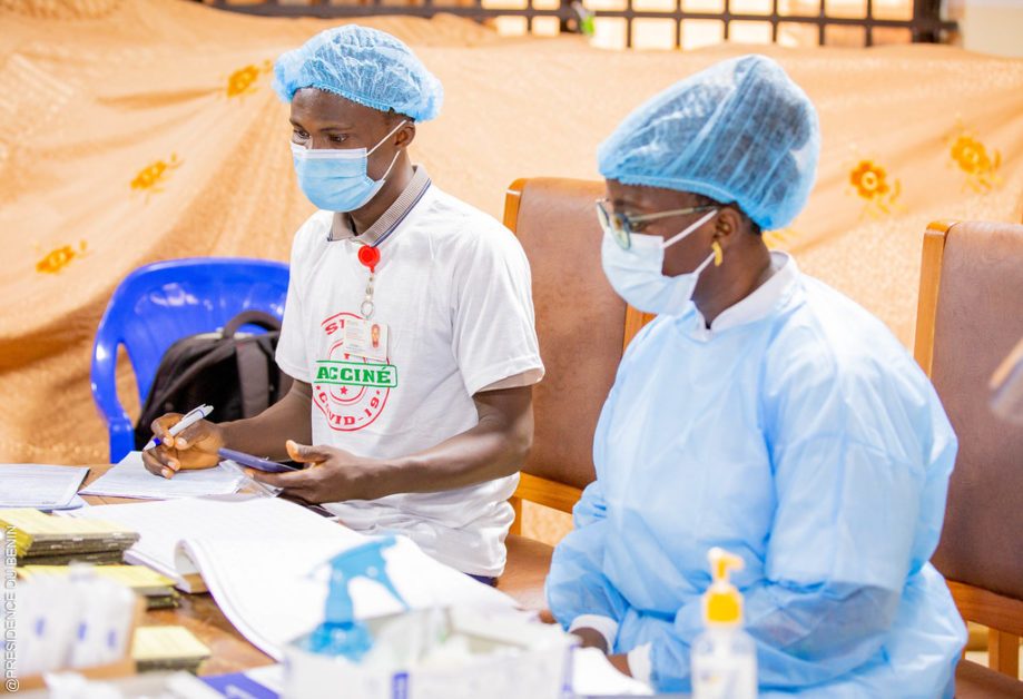 Deux professionnels de santé portant blouse, charlotte et masque sont assis devant un bureau avec documents et flacons sanitaires.