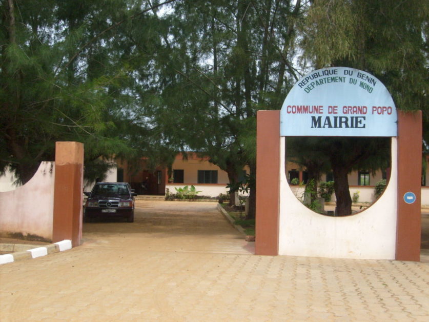 Entrée d un bâtiment administratif en partie masqué par des arbres et un véhicule noir stationné sur une allée beige