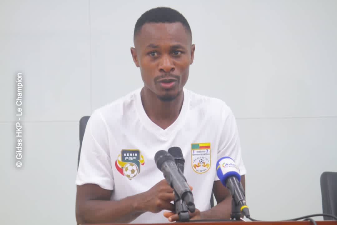 Un homme en polo blanc avec logos de football parle devant deux micros lors d'une conférence de presse intérieure