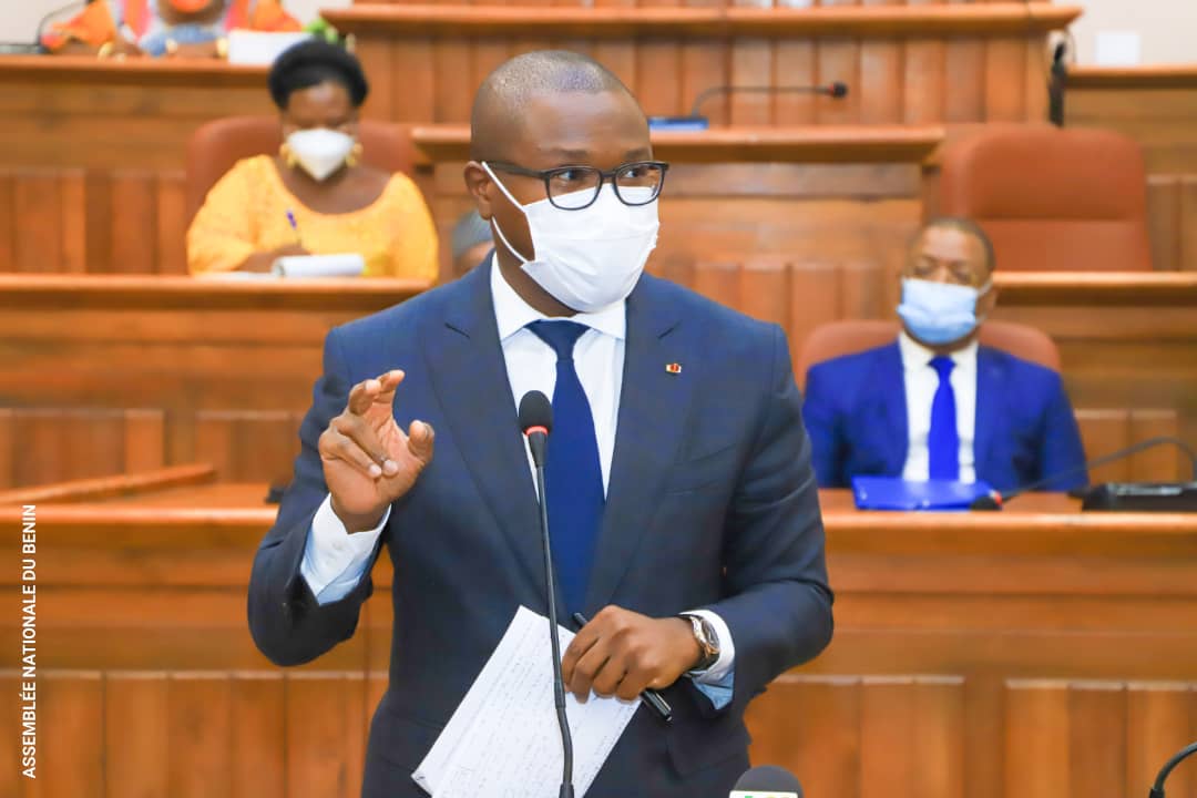 Un homme en costume masque et lunettes parle devant un micro dans une salle d’assemblée avec des personnes assises en arrière-plan