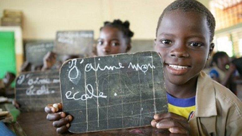 Enfant souriant tenant une ardoise où est écrit j aime mon école dans une salle de classe avec d autres élèves flous en arrière-plan