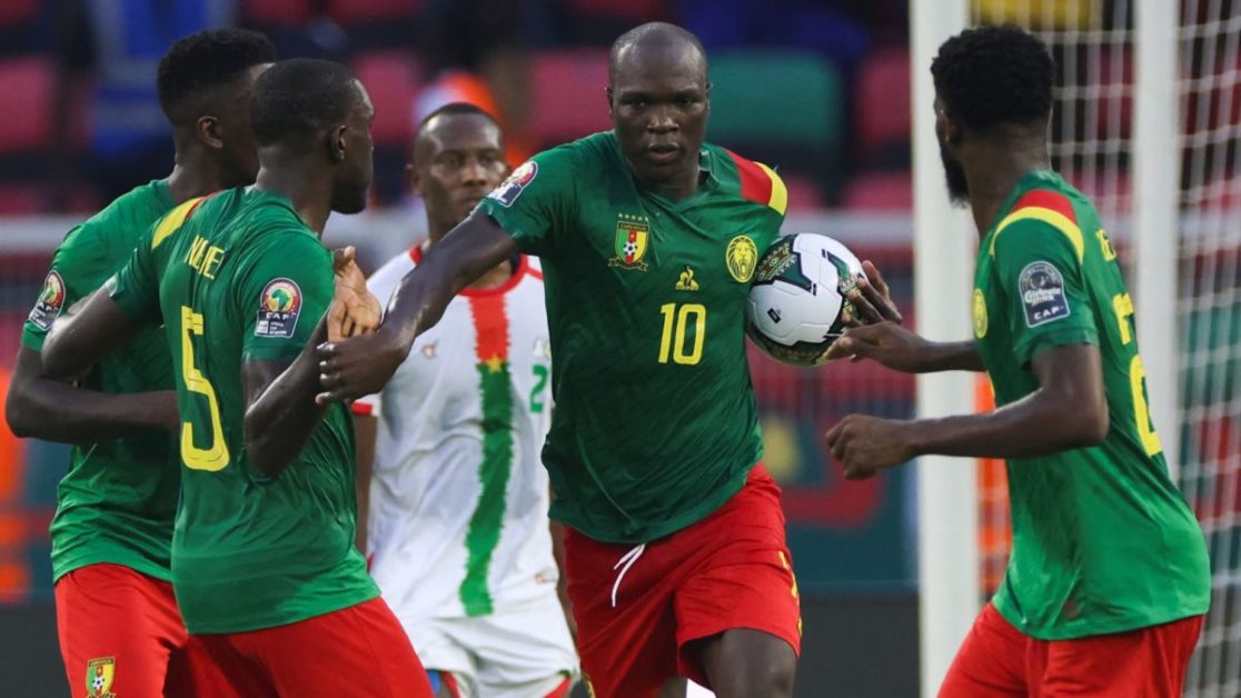 Quatre joueurs en maillots verts et shorts rouges célèbrent un but lors d’un match de football avec un joueur en maillot blanc et vert en arrière-plan