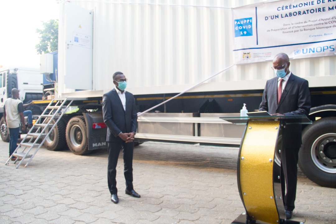 Deux hommes en costume et masque se tiennent devant un laboratoire mobile installé sur un camion blanc