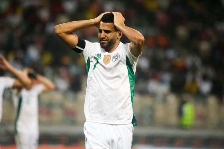 Un joueur de football en maillot blanc et vert se tient la tête à deux mains, visiblement déçu sur le terrain.