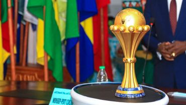 Trophée de football posé sur une table devant plusieurs drapeaux colorés et une personne en costume.