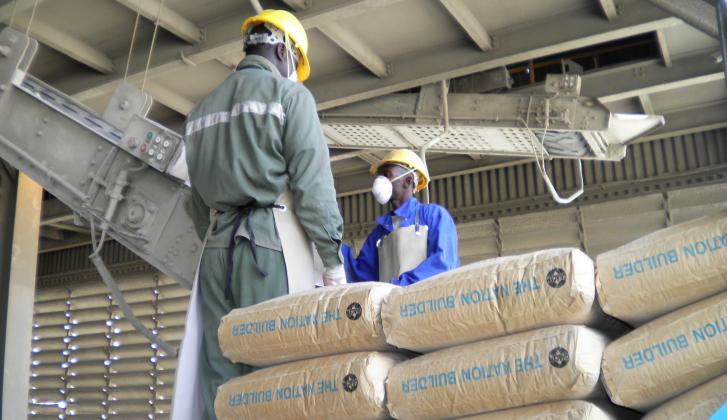 Deux ouvriers en tenue de protection discutent dans une usine avec des sacs de ciment empilés au premier plan