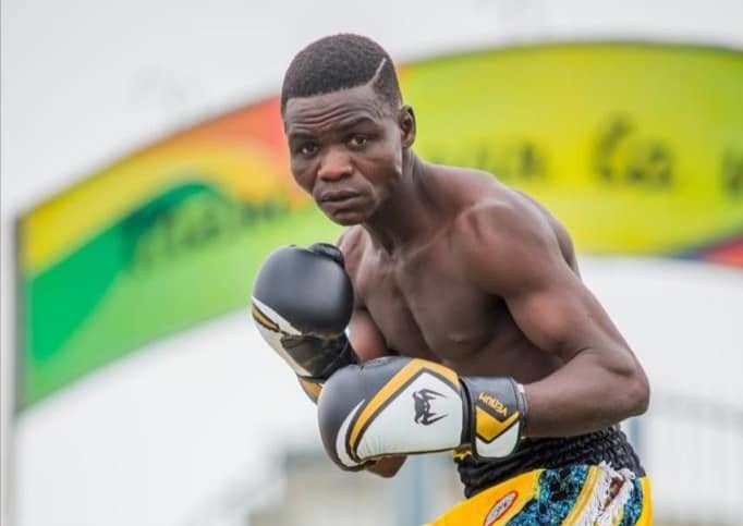 Boxeur torse nu en garde de combat avec gants noirs et jaunes sur un ring en extérieur coloré en arrière-plan