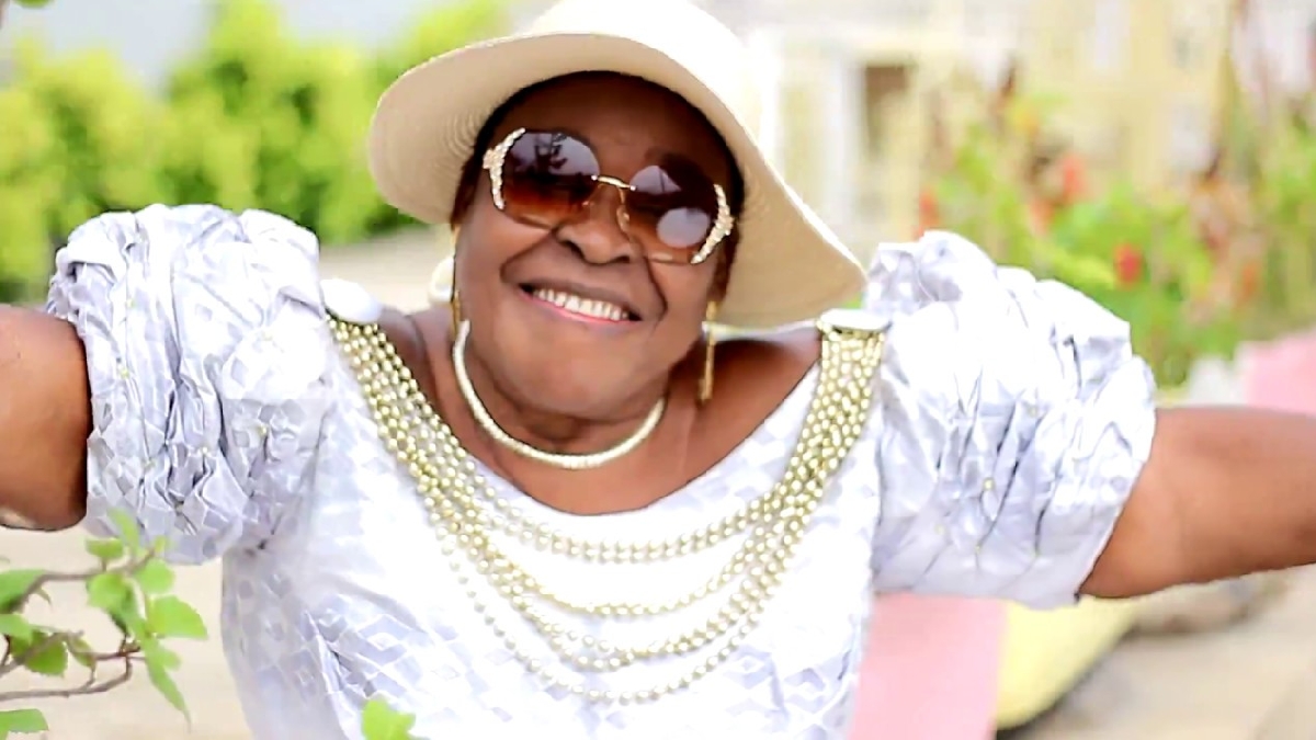 Femme âgée souriante portant des lunettes de soleil, un chapeau beige et une robe blanche ornée de perles, bras ouverts dehors