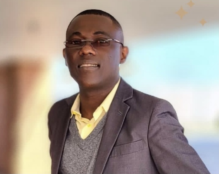 Homme portant des lunettes, un costume sombre avec un gilet gris et une chemise jaune souriant devant un fond flou