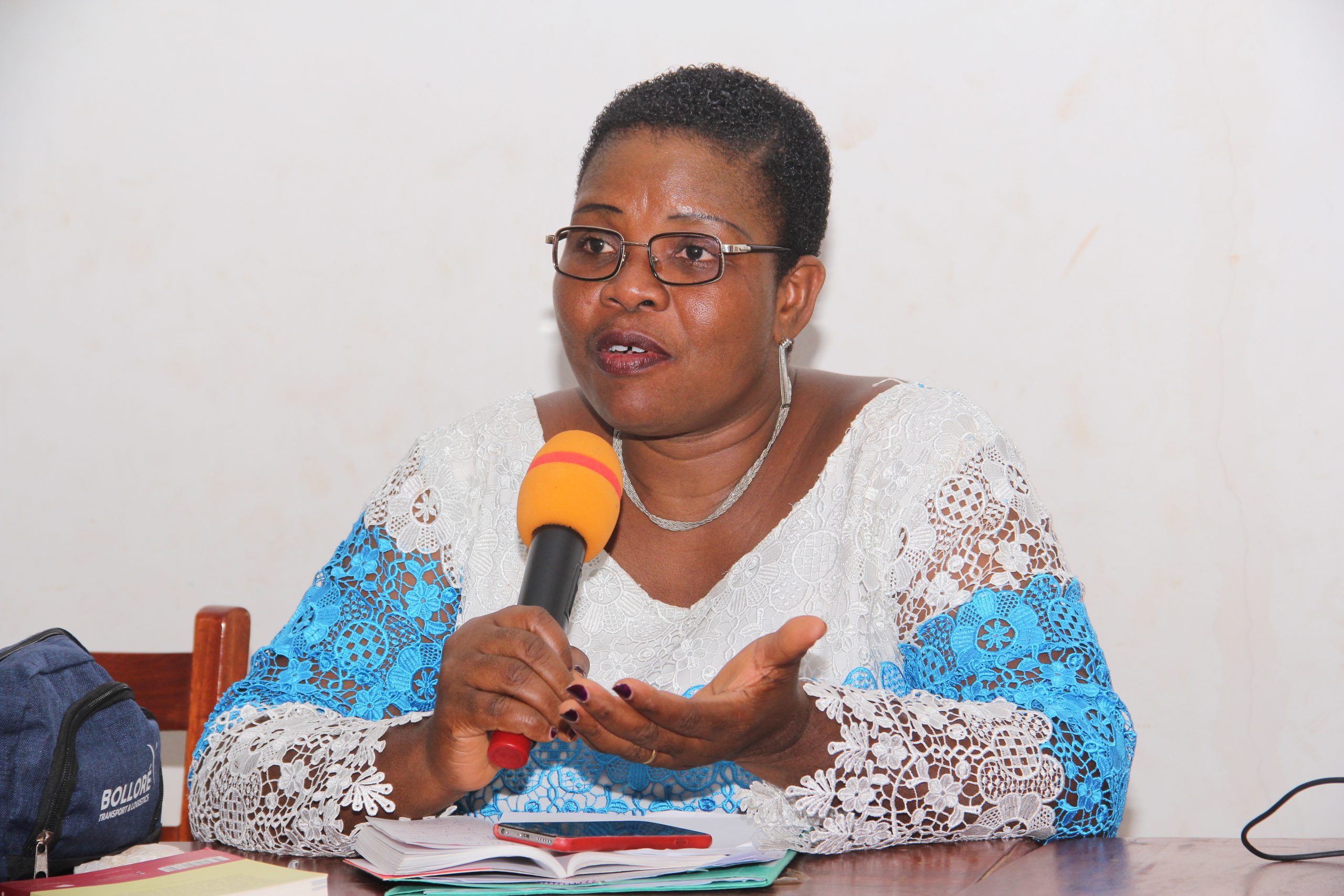 Femme portant des lunettes et une robe en dentelle bleue et blanche parle au micro orange assise devant des documents sur une table