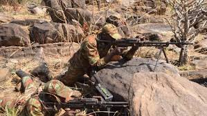 Deux soldats en tenue camouflage armés, positionnés derrière des rochers dans un environnement sec et rocailleux