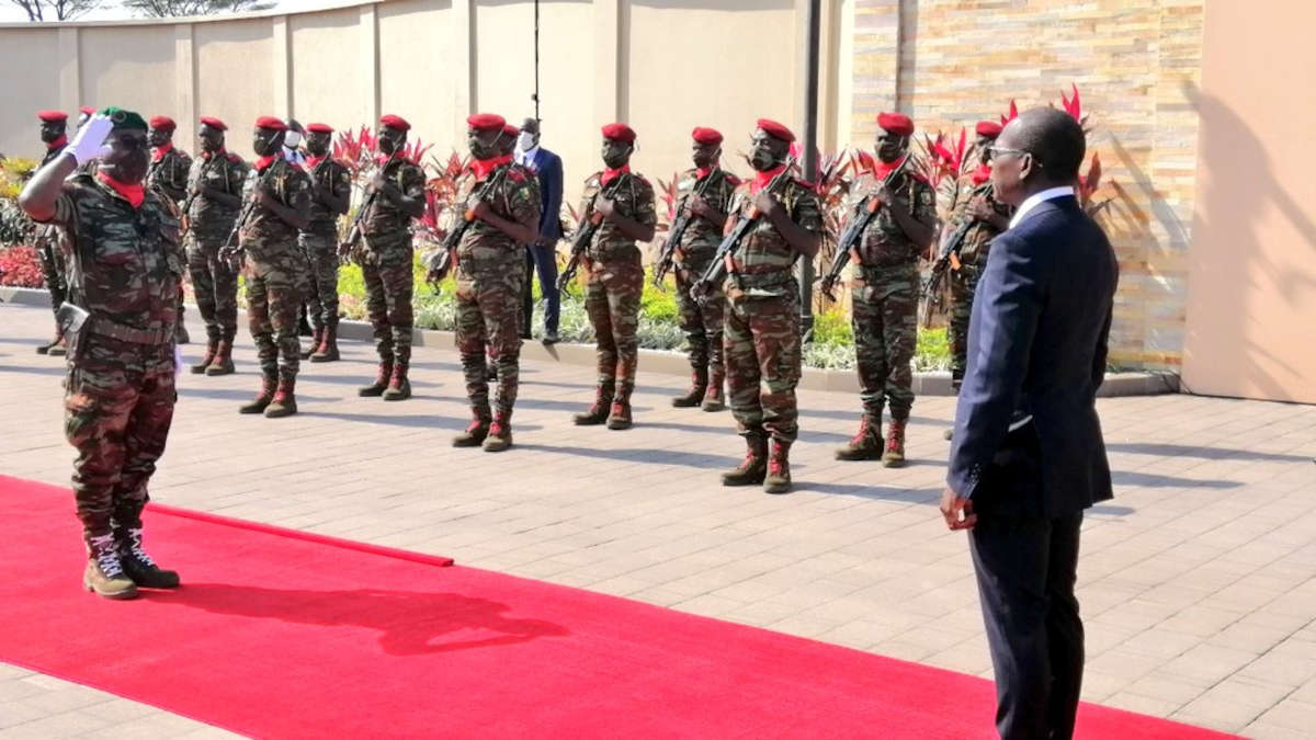 Un groupe de soldats en tenue camouflée et bérets rouges en formation face à un homme en costume sur un tapis rouge devant un mur beige