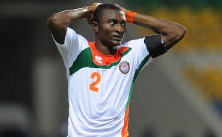 Joueur de football en maillot blanc aux bandes vertes et oranges les mains sur la tête expression contrariée sur le terrain