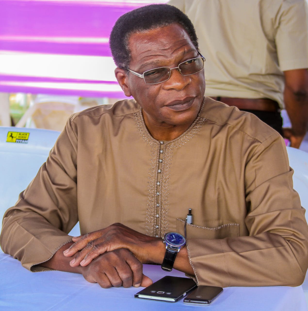 Homme portant une tunique beige et des lunettes assis à une table avec deux téléphones posés devant lui