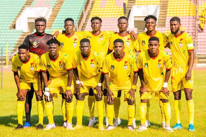 Onze jeunes hommes en tenue de football jaune posent en rang devant un stade aux gradins colorés