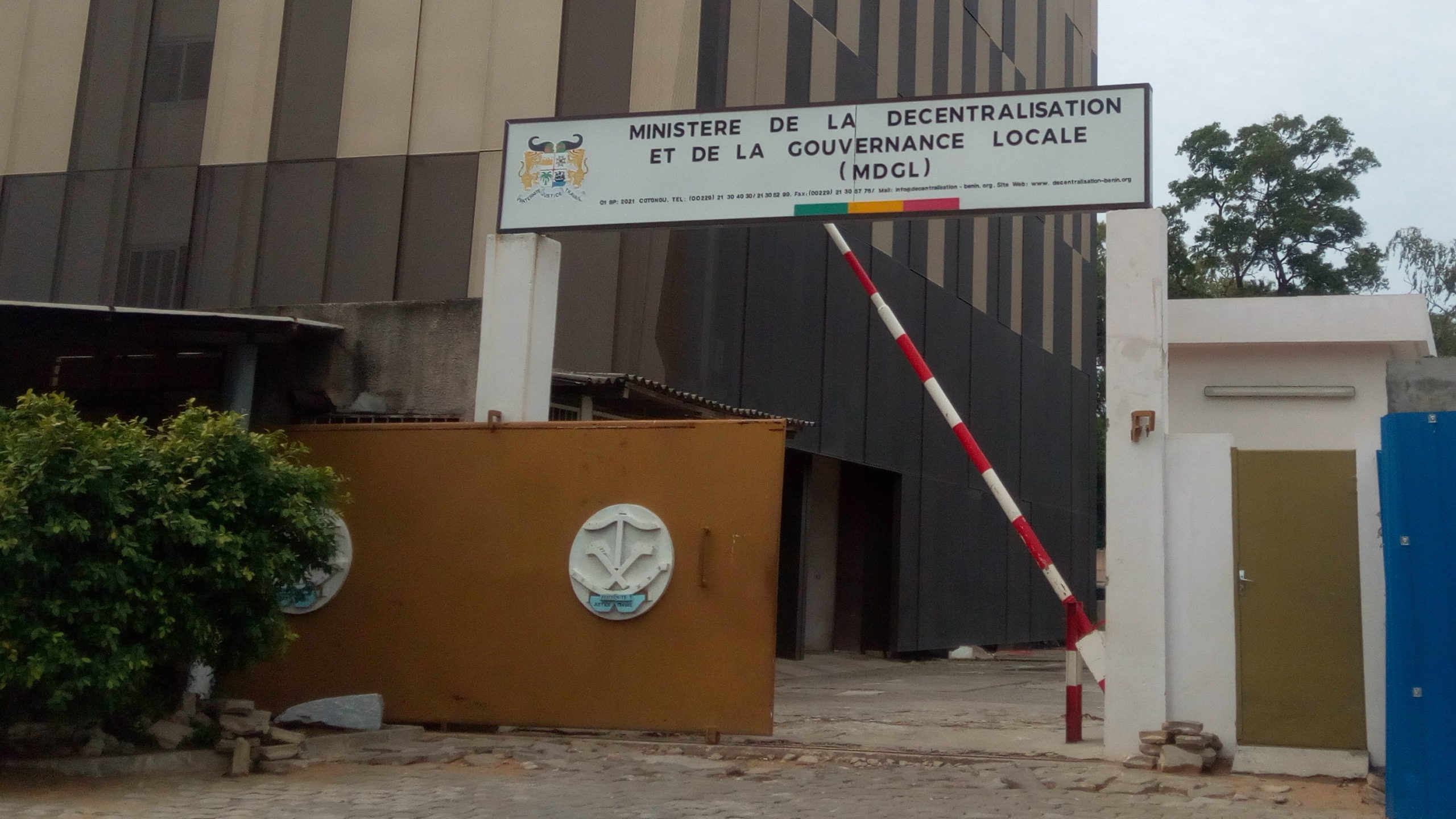 Entrée d’un bâtiment administratif avec un panneau du ministère de la décentralisation et de la gouvernance locale suspendu au-dessus d’une barrière rouge et bl
