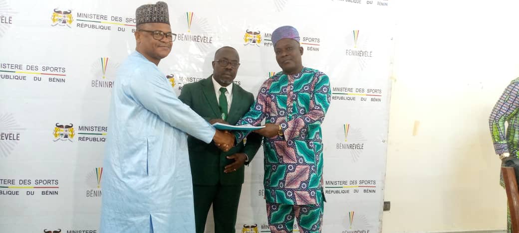 Trois hommes en costume traditionnel et costume formel se serrent la main devant un mur avec logos officiels liés au sport du Bénin
