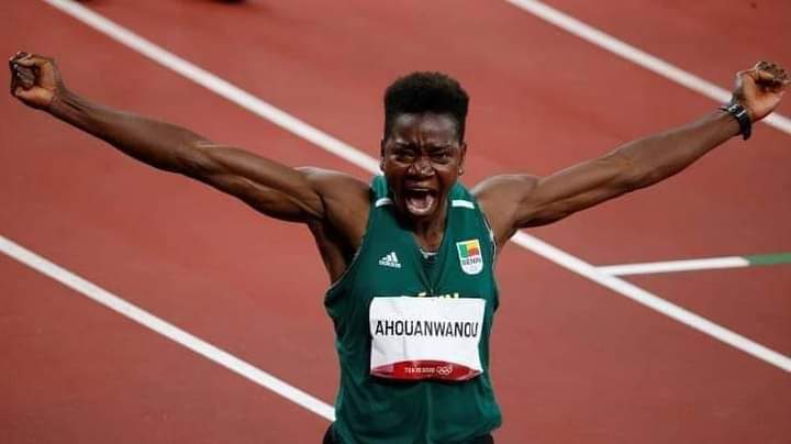 Une athlète en tenue verte célèbre en levant les bras sur une piste d’athlétisme rouge