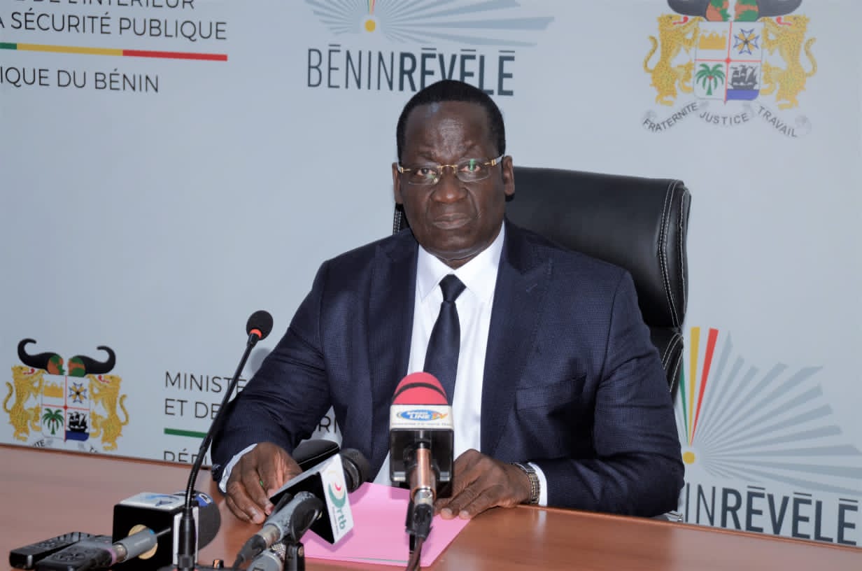 Homme en costume sombre assis à une table avec plusieurs microphones devant lui dans une salle officielle