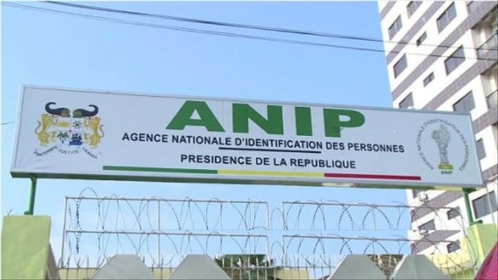 Panneau avec le sigle ANIP Agence Nationale d’Identification des Personnes au-dessus d’une clôture et de barbelés devant un immeuble.