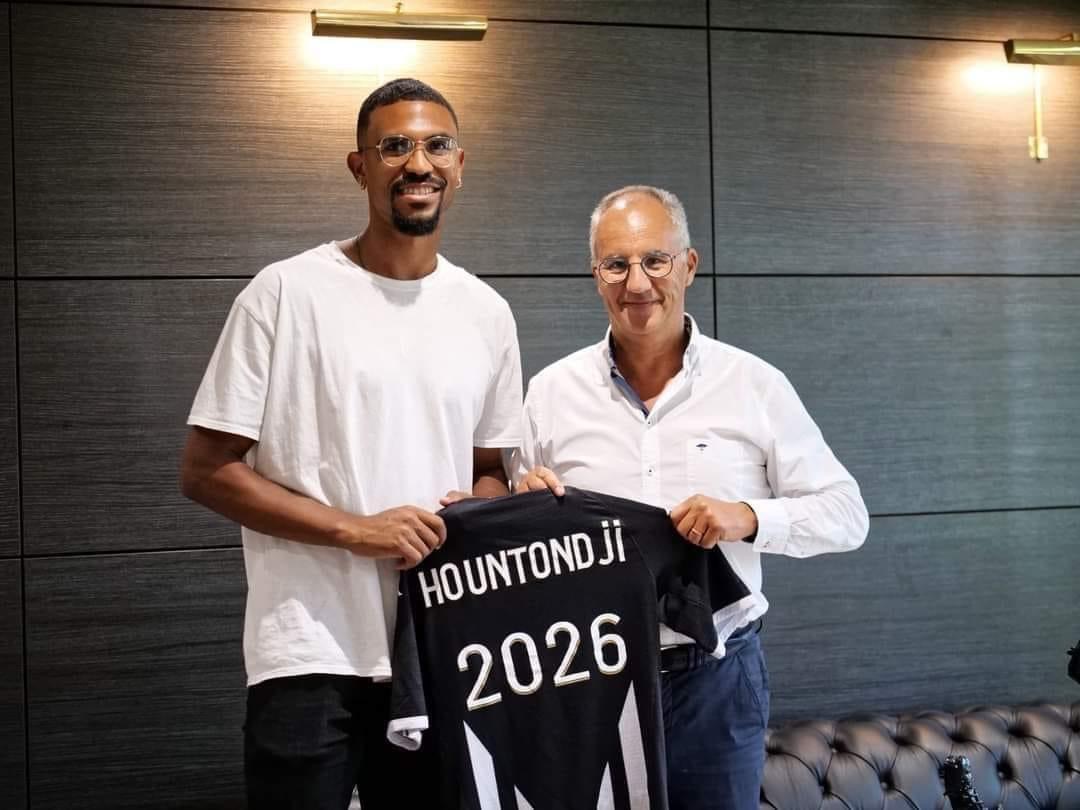 Deux hommes souriants tiennent un maillot de football noir avec le nom Hountondji et l'année 2026 sur un fond de mur gris