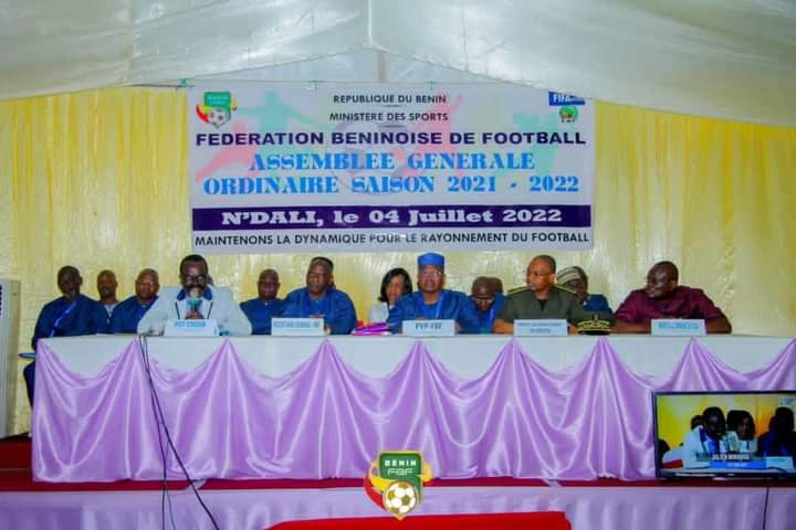 Plusieurs personnes assises derrière une table drapée de tissu violet sous une banderole de l'assemblée générale de football 2021-2022