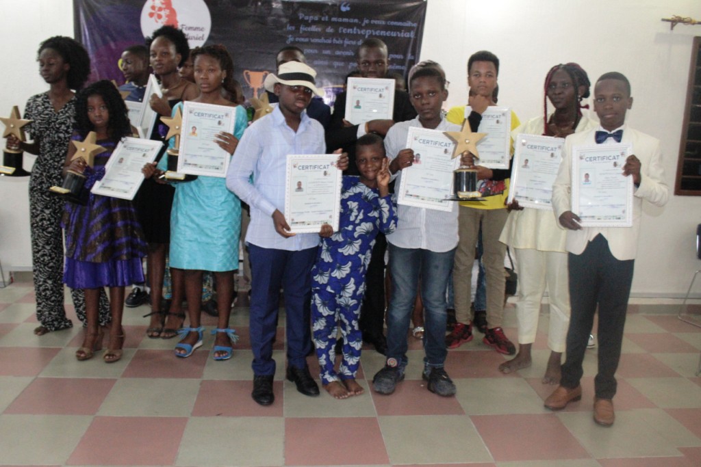 Un groupe d’enfants posant debout, tenant des certificats et des trophées en forme d’étoile dans une salle.