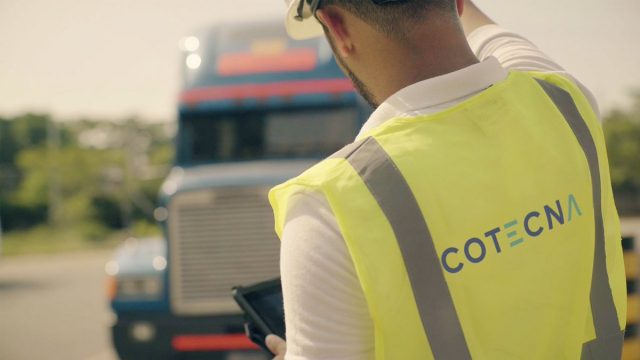 Homme en gilet jaune avec inscription COTECNA utilisant une tablette devant un camion bleu flou en arrière-plan