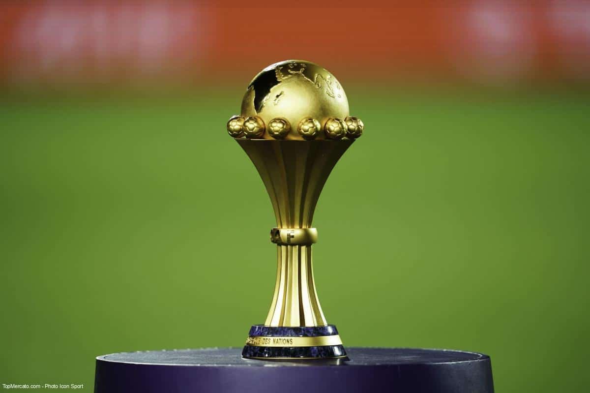 Trophée doré de la Coupe d'Afrique des Nations posé sur un socle contre un fond flou vert et rouge