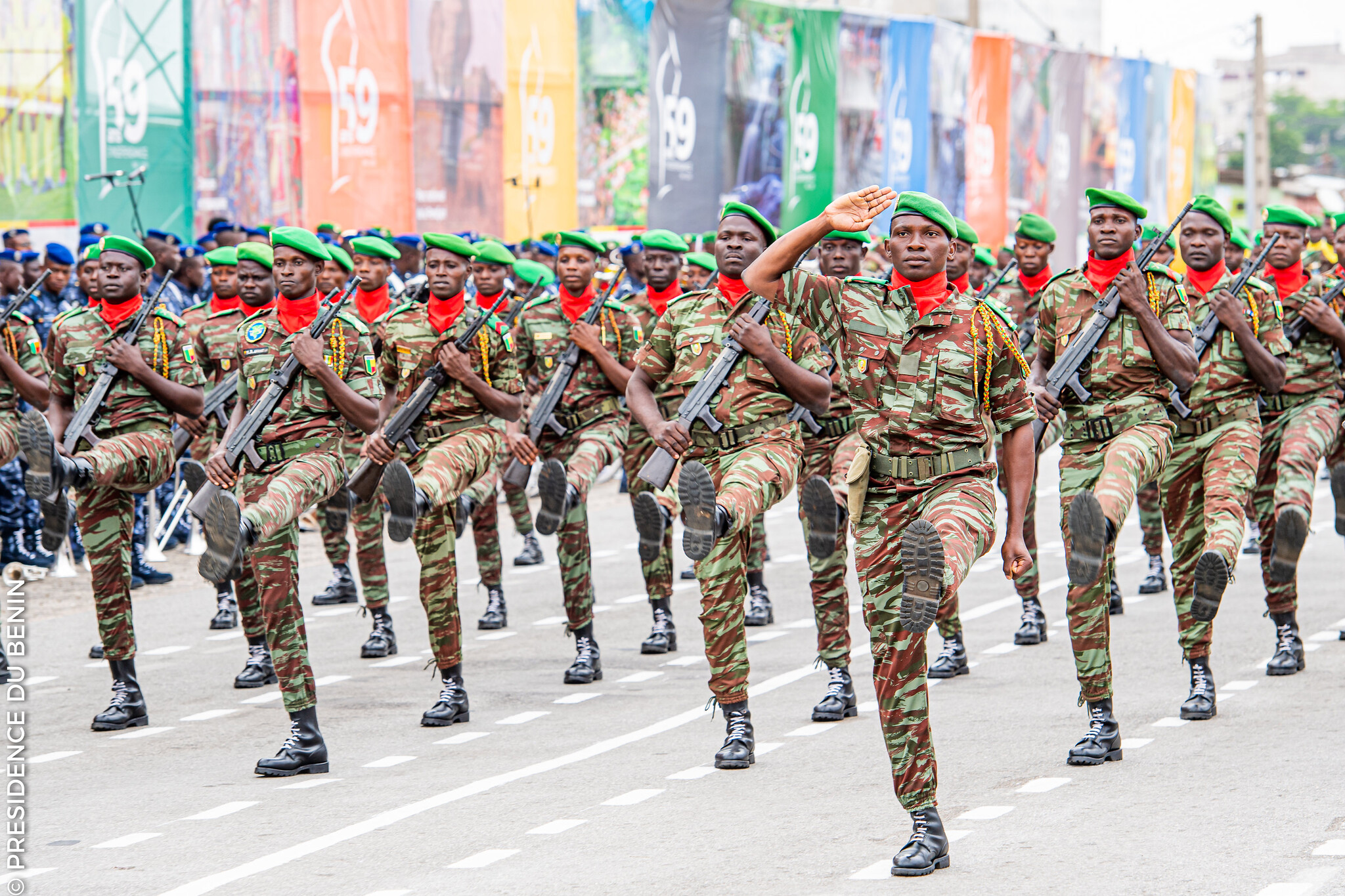 Des soldats en uniforme camouflage et bérets verts défilent en formation serrée sur une avenue bordée de drapeaux colorés