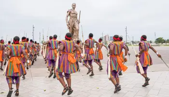 Groupe d’hommes en tenues traditionnelles colorées dansant devant une statue monumentale sur une place ouverte