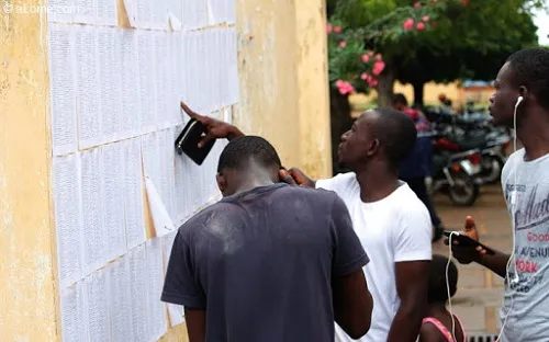 Quatre jeunes hommes consultent une liste affichée sur un mur extérieur sous un arbre avec des motos en arrière-plan