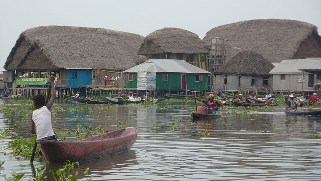 Personne pagaie sur une pirogue dans un village lacustre avec maisons sur pilotis et toits de chaume