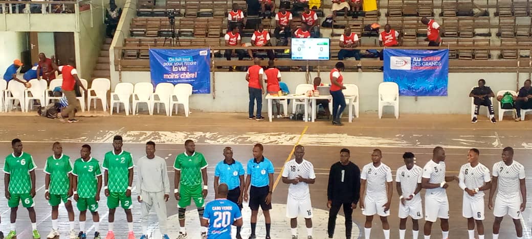 Deux équipes de handball en rang face à face avec deux arbitres au centre dans une salle avec tribunes et spectateurs