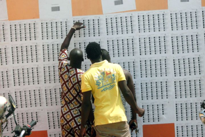 Trois hommes debout lisent attentivement une grande liste affichée sur un mur orange et blanc