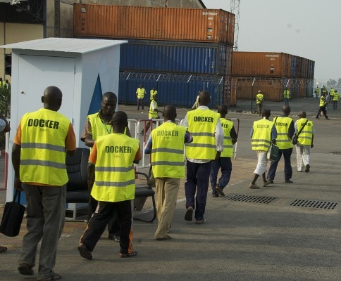 Des hommes portant des gilets fluorescents marqués « DOCKER » marchent près de conteneurs empilés sur un quai portuaire