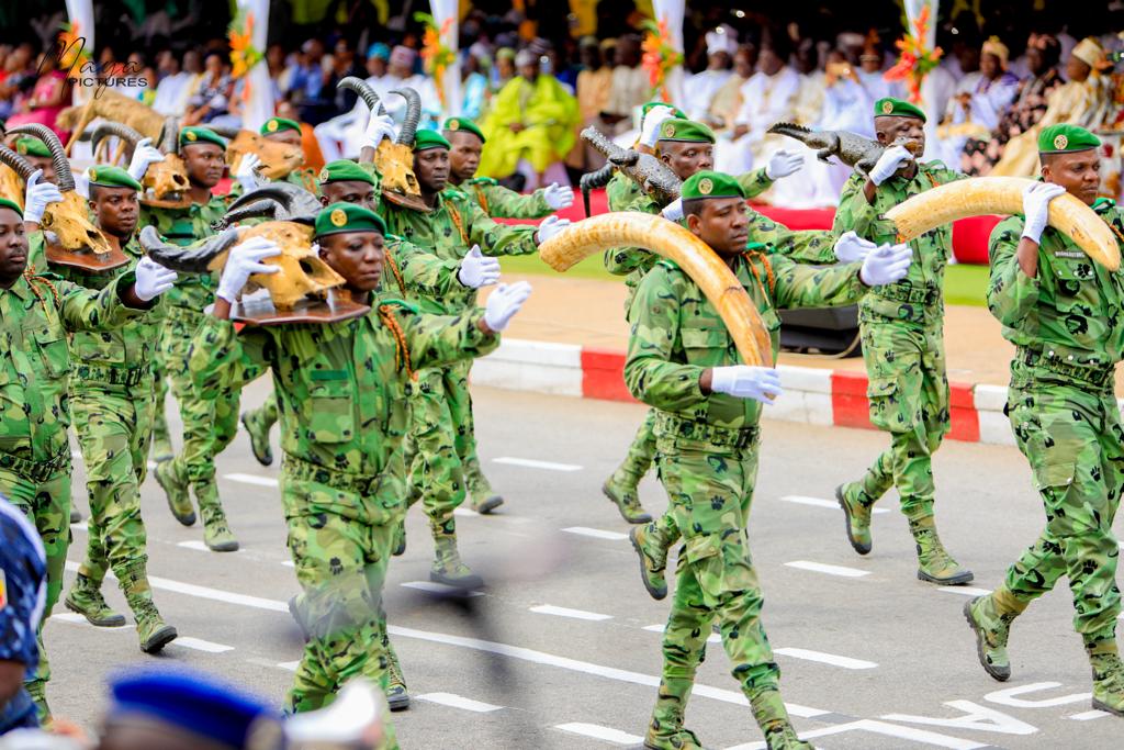 Des militaires en uniforme camouflage vert défilant, certains portant des têtes et des défenses d’animaux devant un public assis