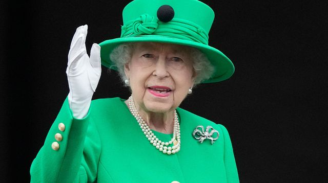 Personne âgée souriante en manteau vert avec chapeau assorti, gant blanc, collier de perles et broche, saluant de la main droite face caméra