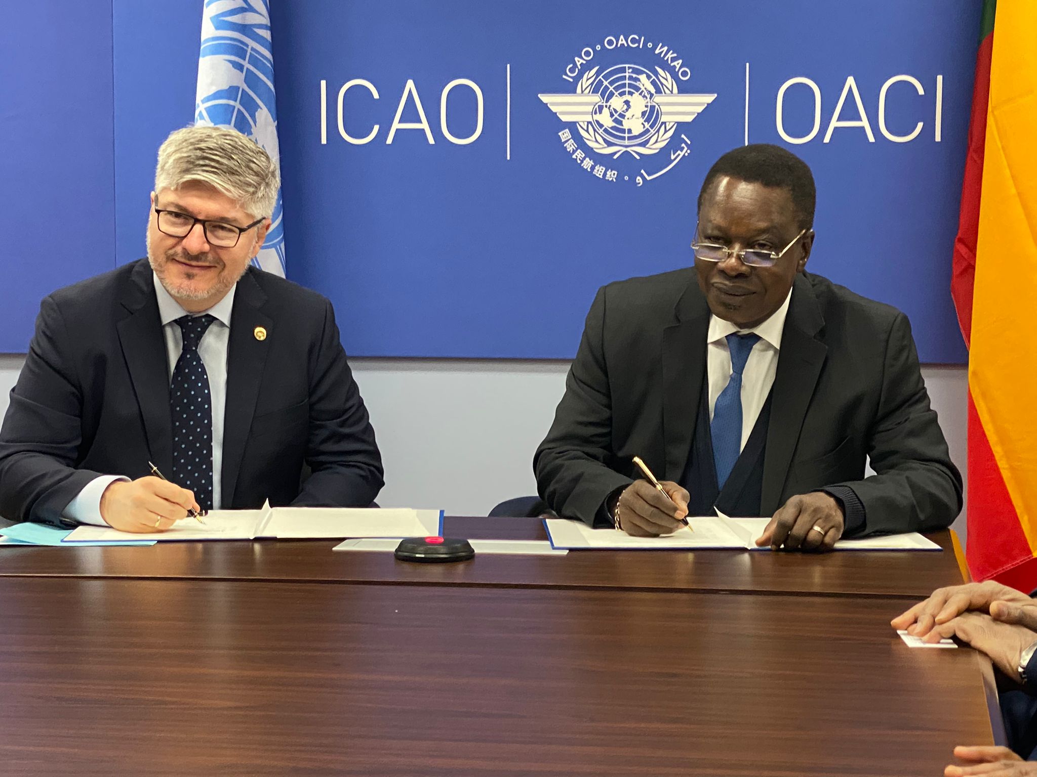 Deux hommes en costume signent des documents devant un panneau bleu marqué ICAO OACI avec un drapeau partiellement visible à droite