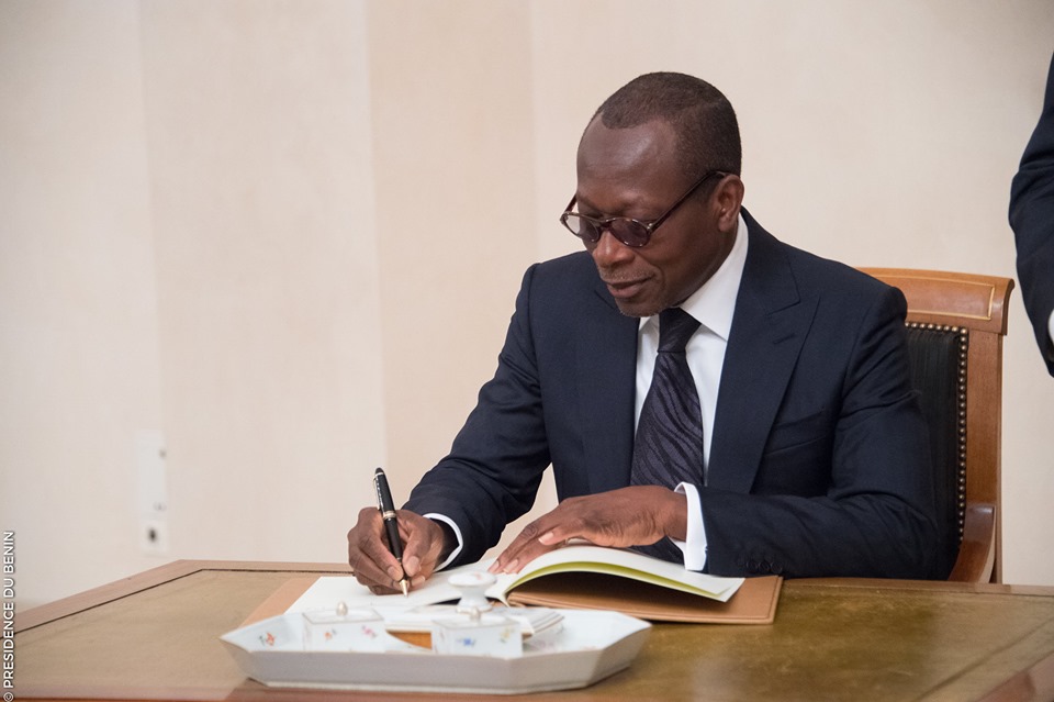 Un homme en costume sombre et lunettes signe un document posé sur une table en bois clair devant un mur beige
