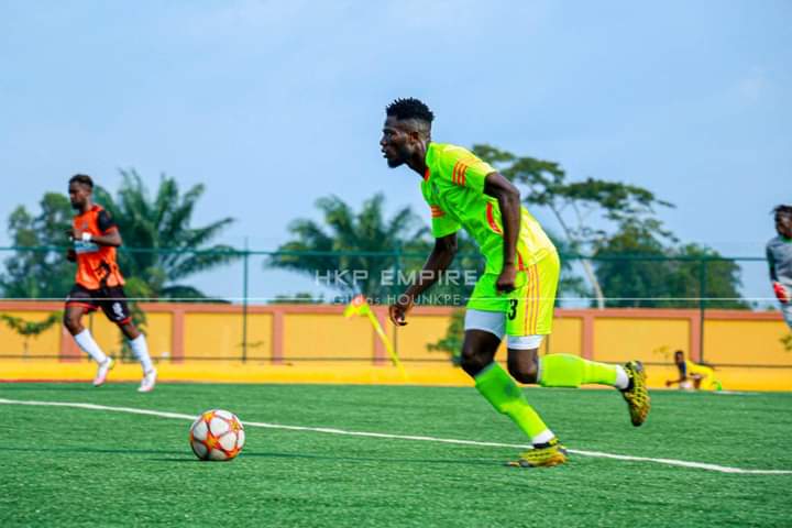 Joueur de football en tenue verte courant vers le ballon sur un terrain synthétique entouré d'autres joueurs en arrière-plan