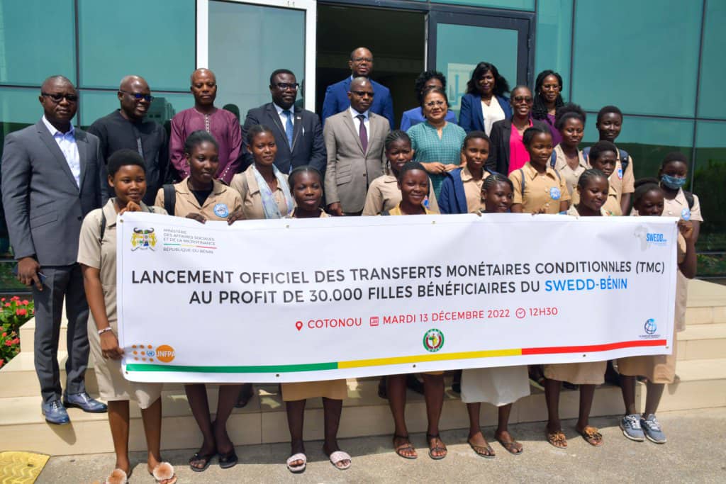 Un groupe d’adultes et d’adolescentes posent devant un bâtiment avec une grande bannière annonçant un lancement officiel de transferts monétaires conditionnels