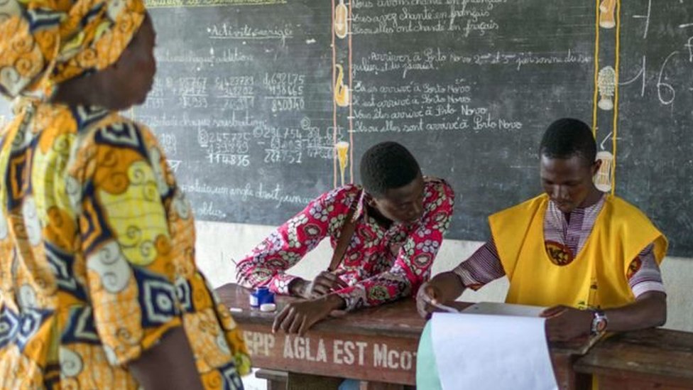 Deux personnes assises à une table vérifient des documents face à une femme debout dans une salle avec un tableau noir en arrière-plan